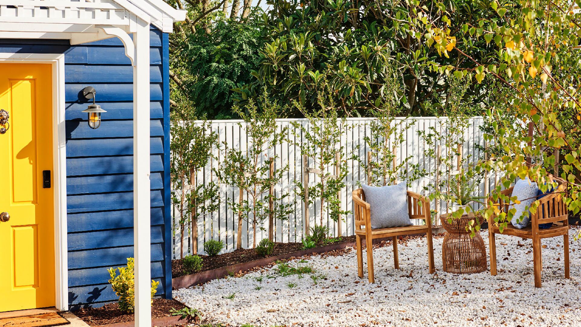 A blue shed with a yellow door, a white fence, and a seating area with two wooden chairs and a wicker basket.