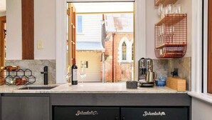 A kitchen with a marble countertop, a wine bottle, and a wine glass. There is a red wire basket on the wall and a wooden shelf with glasses.