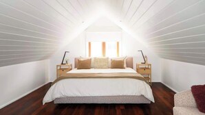 A bedroom with a sloped ceiling, a large bed with a white duvet, two bedside tables with lamps, and a window with curtains.