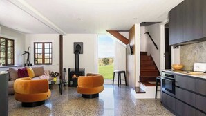 A modern living room with a fireplace, a sofa, and a kitchen area with dark cabinetry and a wooden staircase.