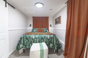 A bedroom with a bed, a bench, a window with curtains, a painting on the wall, and a ceiling light.