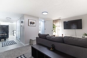 Uma sala de estar moderna com um sofá, uma TV de tela plana, um espaço de jantar com mesa e cadeiras, e um corredor que leva a outro cômodo.