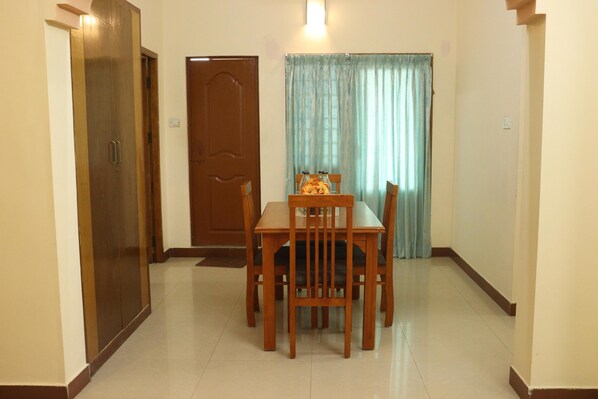A dining area with a wooden table and chairs, a door, and a window with curtains. - Le Ideal Suites (Puducherry)
