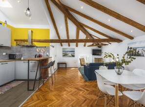 Une cuisine moderne avec un mur d’accent jaune, des poutres en bois et un coin repas comprenant une table et des chaises blanches.