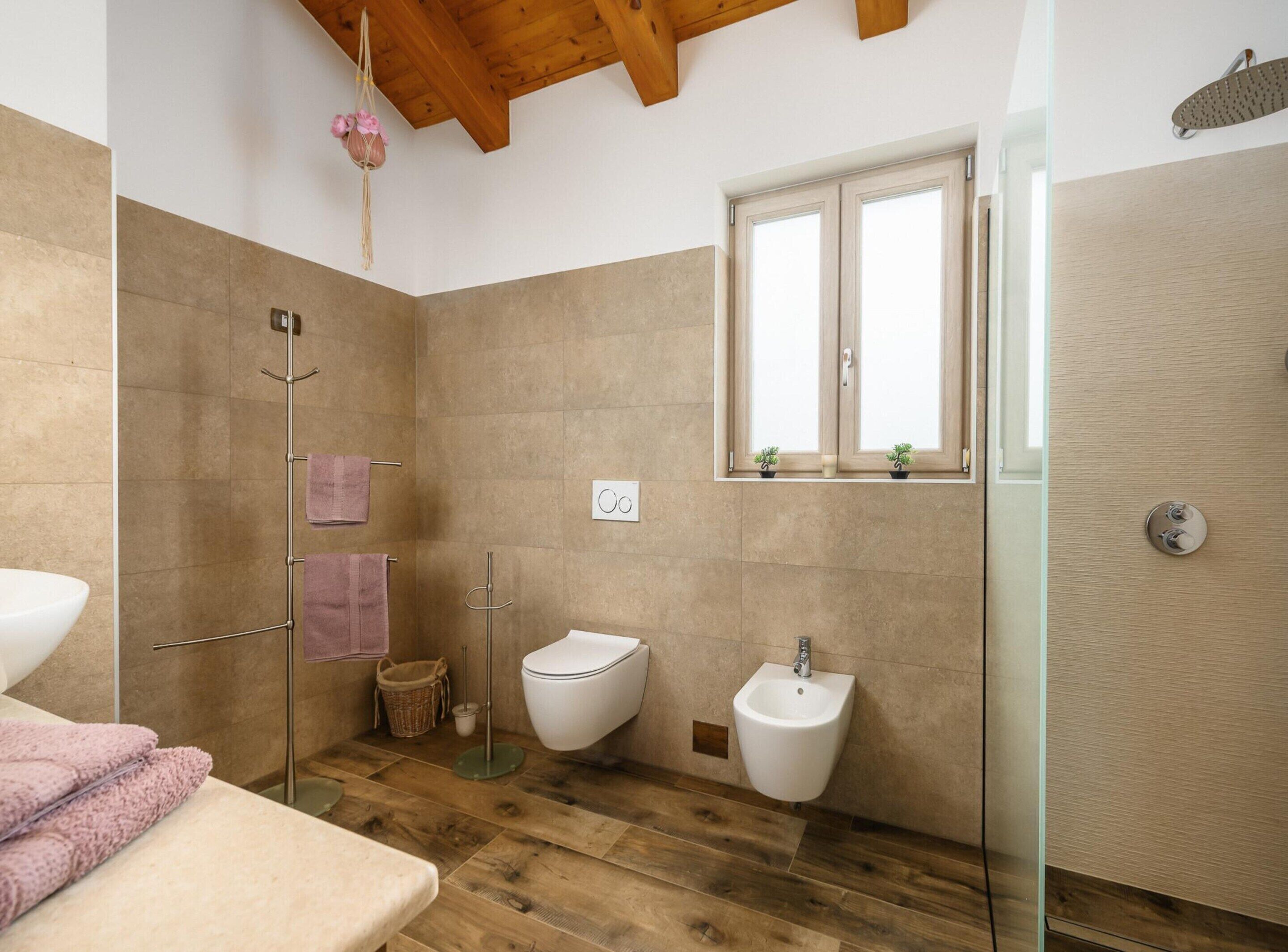 A modern bathroom with a wooden floor, a glass shower enclosure, a bidet, and a window with plants.
