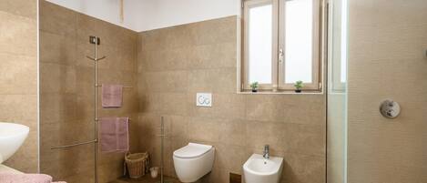 A modern bathroom with a wooden floor, a glass shower enclosure, a bidet, and a window with plants.