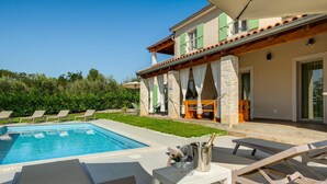 Un espace aménagé au bord de la piscine, avec des chaises longues, une table sur laquelle est posée une bouteille de champagne, et un bâtiment en pierre doté d’un balcon.