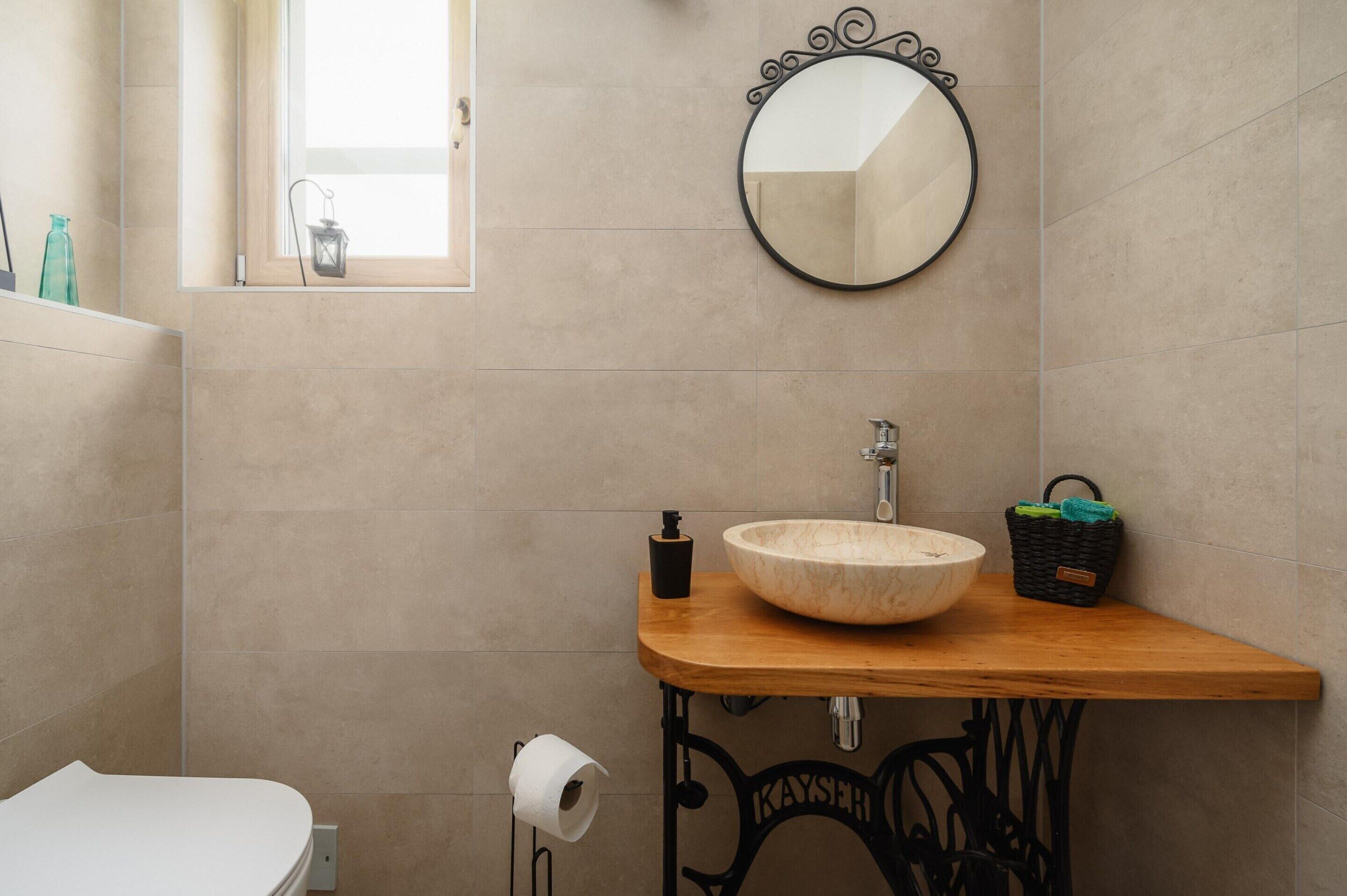 A bathroom with a wooden vanity, a round mirror, a sink, and a window with a view.