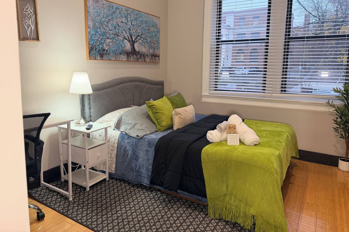 A neatly arranged bedroom with a bed, bedside table, lamp, and a window with blinds.