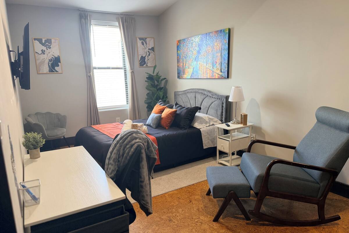 A bedroom with a bed, bedside table, chair, desk, and a painting on the wall.