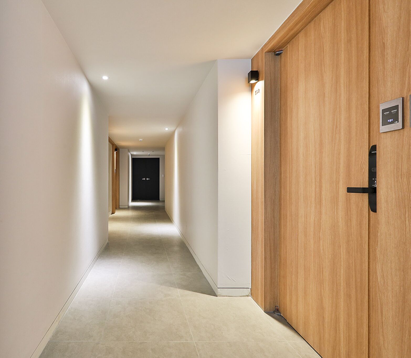A modern hotel corridor with a wooden door on the right, a light fixture above it, and a doorbell.