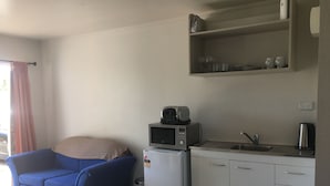 A compact kitchenette with a microwave, toaster, and sink. There is a blue chair and a small trash bin.