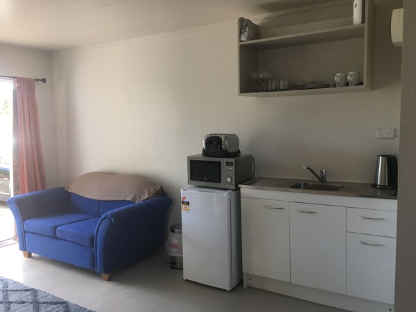 Una kitchenette compacta con microondas, tostadora y lavamanos. Hay una silla azul y un pequeño cubo de basura.