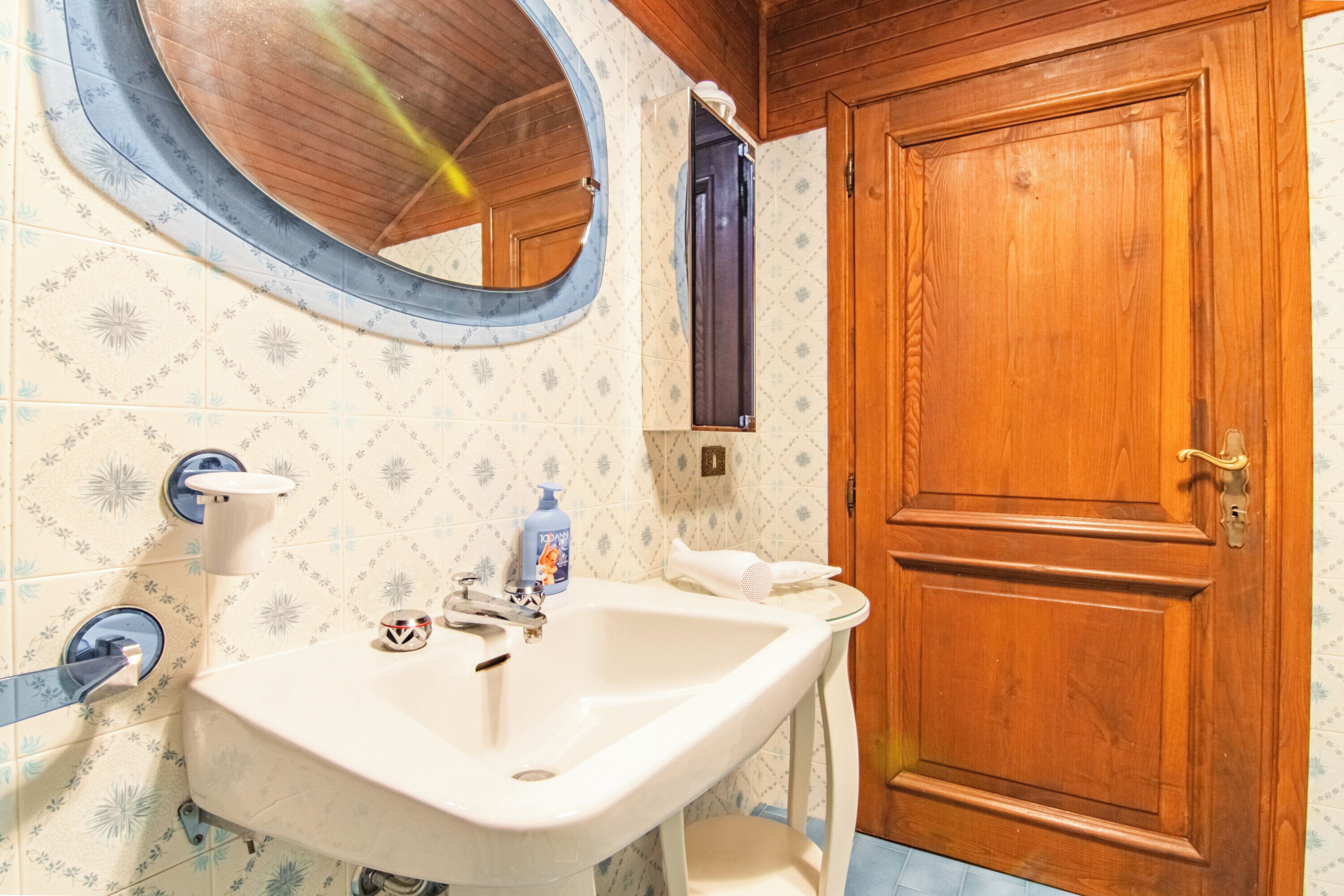 Un baño con puerta de madera, un espejo redondo, un lavamanos y azulejos estampados.