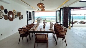 A spacious dining area with a long table and wicker chairs, large windows offering a view of the landscape, and decorative wall art. - Villa Teraum (Kuta)