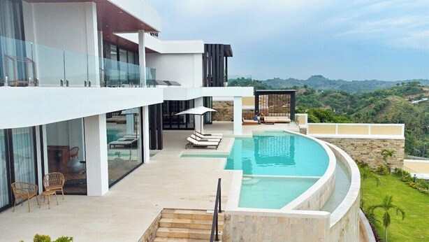 A modern two-story building with a curved infinity pool, outdoor seating, and a view of the hills. - Villa Teraum (Kuta)