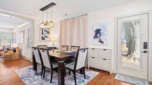 Et moderne spiseområde med et træbord, seks stole og en lysekrone. Der er et stort vindue med udsigt til et træ og en dør med en kodeadgangslås.