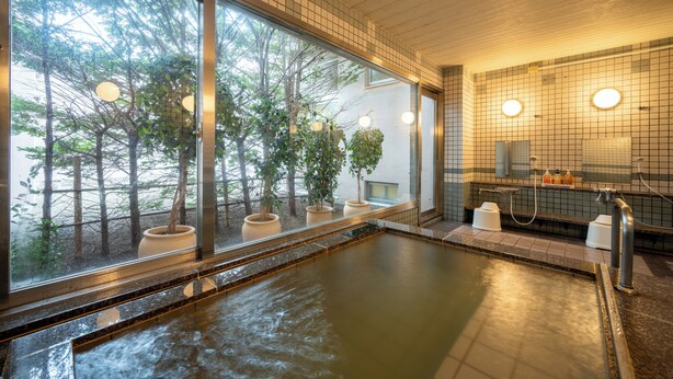 Baño de aguas termales con vistas a árboles a través de amplios ventanales, techo de madera y pared con azulejos, espejo y lavamanos.