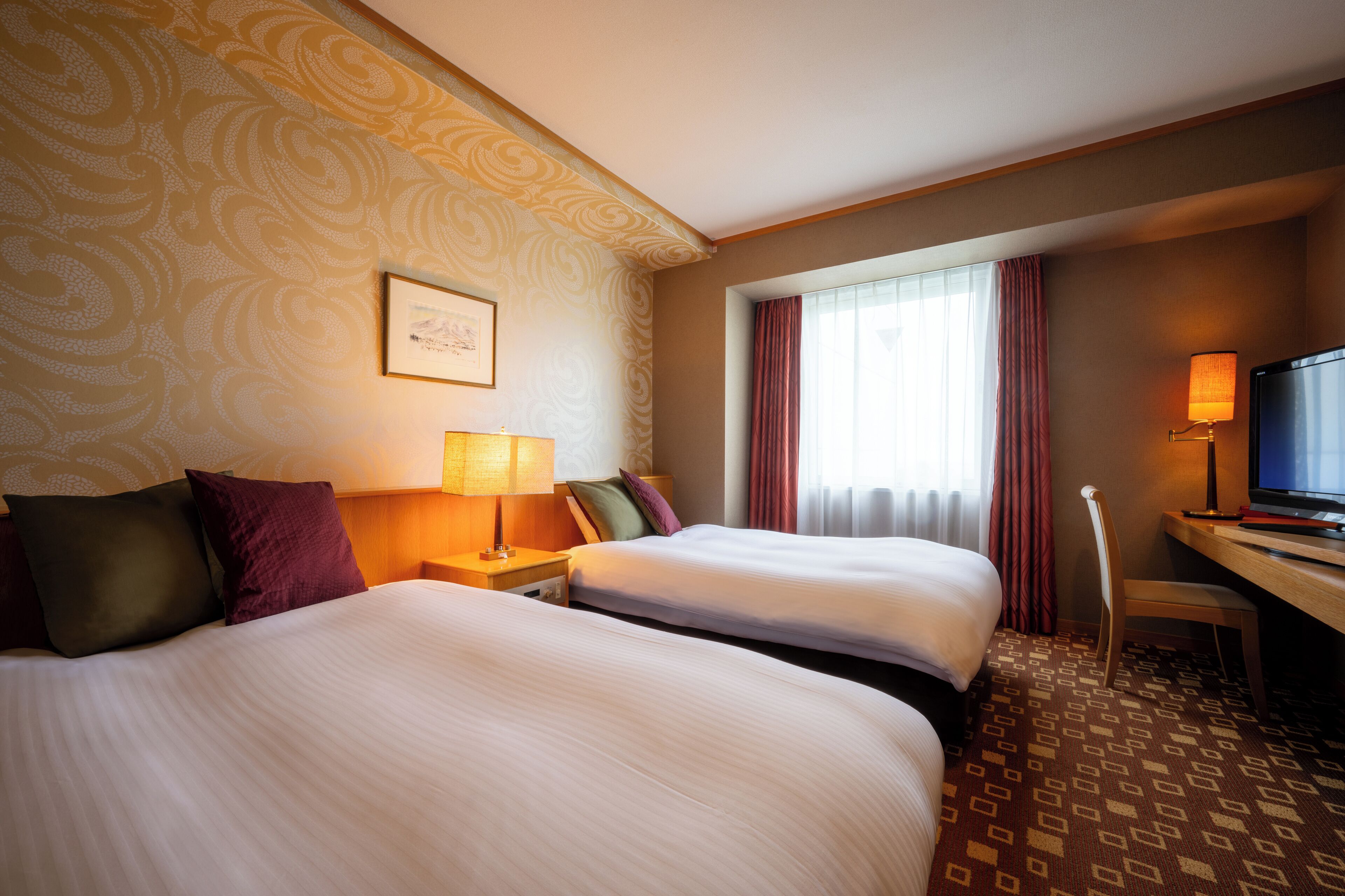 A hotel room with two beds, a desk, a chair, a TV, and a window with curtains.