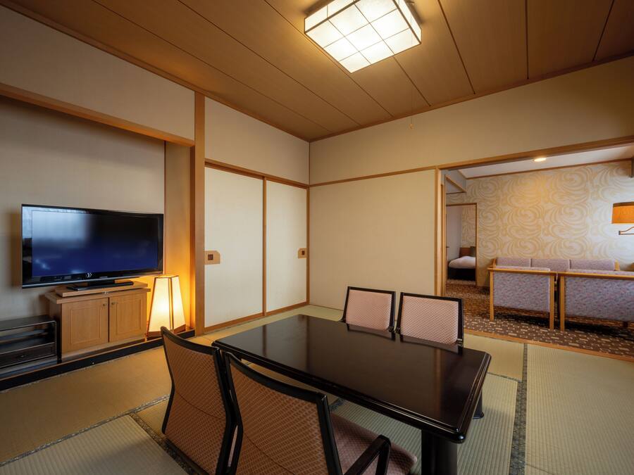 Ein traditioneller japanischer Raum mit Tatami-Boden, einem niedrigen Tisch mit Stühlen, einem Fernseher und einer Couch.