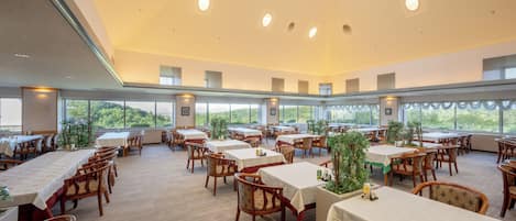A spacious dining area with multiple tables and chairs, large windows offering a view of greenery, and a high ceiling with recessed lighting.