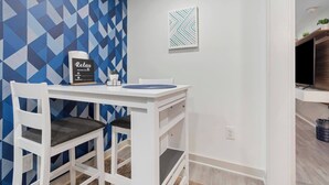 A small dining area with a white table and chairs, a geometric-patterned wall, a framed sign, and a small plant on a shelf. - Marietta Square Suites - Suite3 - Cozy Studio! (Marietta)
