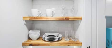 A kitchen with white tiled walls, wooden shelves holding plates, bowls, and wine glasses.
