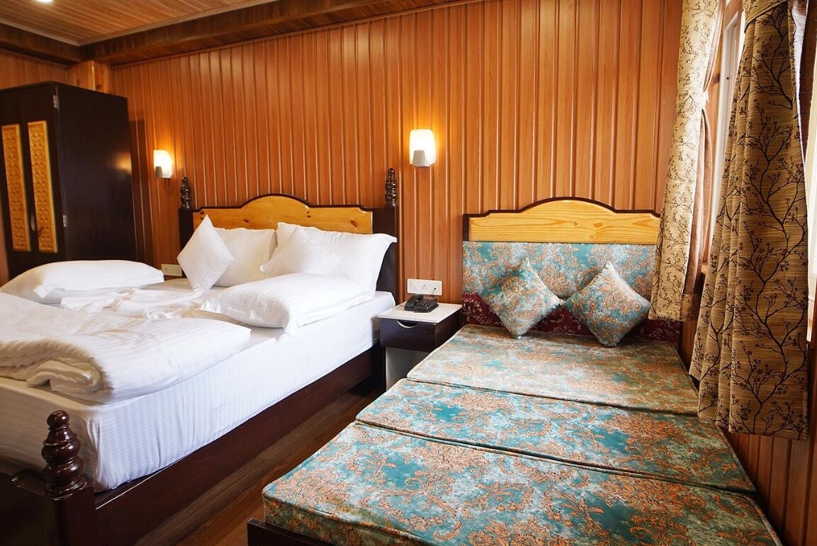 A hotel room with a wooden paneled wall, a large bed with white linens, a bedside table with a lamp, and a patterned bench.
