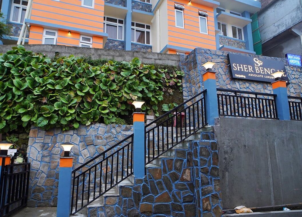 A hotel entrance with a stone staircase, a sign reading "Sher Ben Cial," and a building with orange and blue exterior walls.