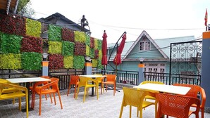 Een terras met kleurrijke stoelen en tafels, een decoratieve muur met planten, en een traditioneel gebouw op de achtergrond.