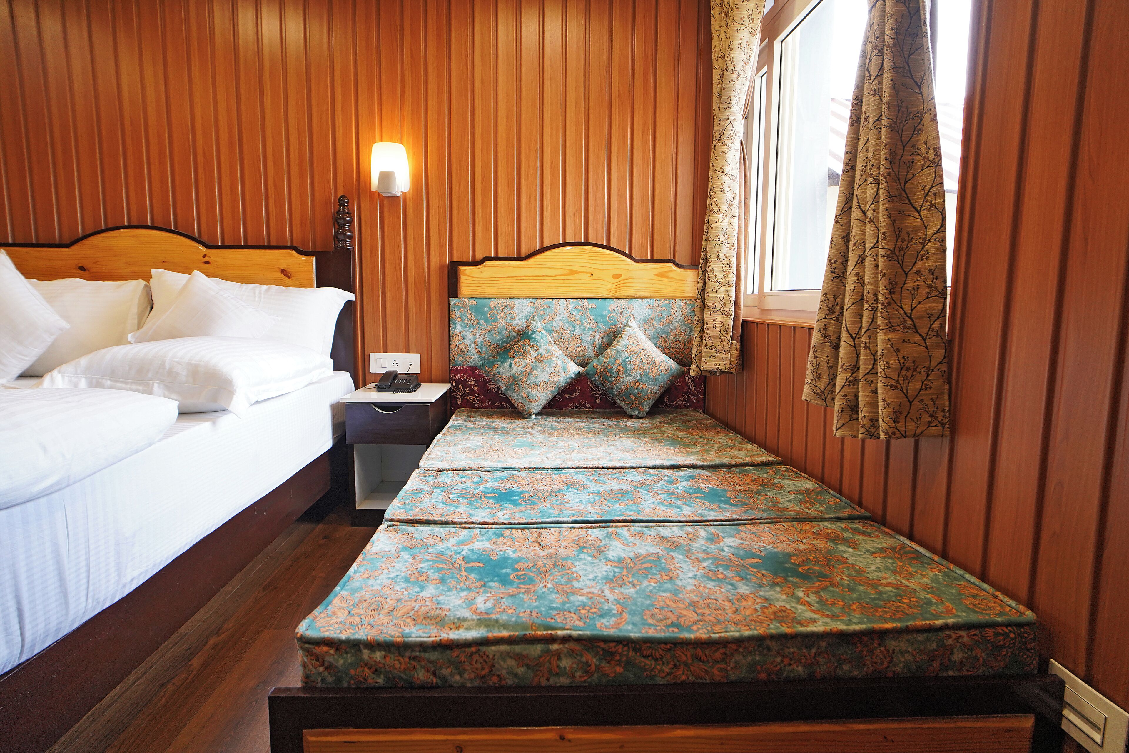 A room with a wooden paneled wall, a bed with white bedding, a bench with patterned cushions, a nightstand, and a window with curtains.