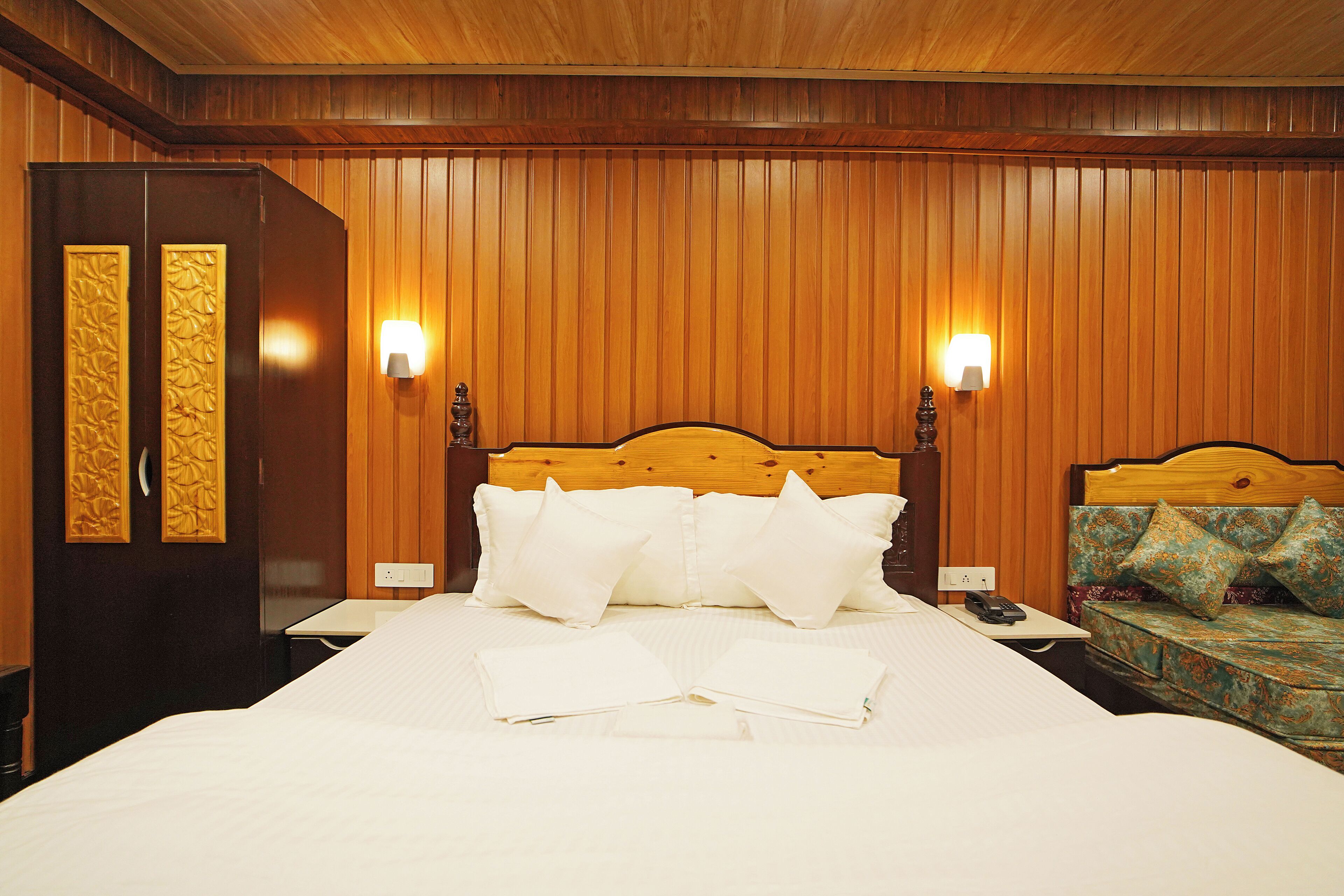 A hotel room with a wooden paneled wall, a large bed with white linens, two bedside lamps, and a nightstand with a phone.