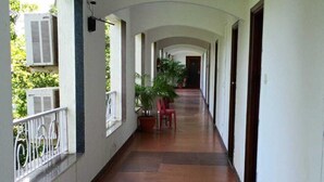 A long corridor with tiled flooring, white walls, and multiple doors. There are potted plants and a red chair along the corridor. - Arnala Beach Resort (Vasai)