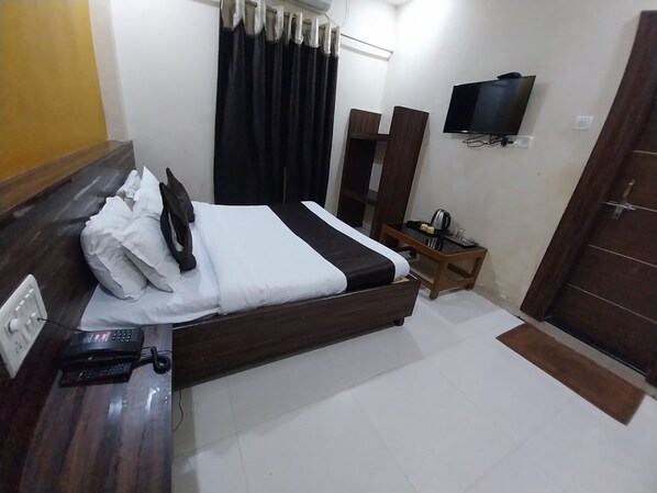 A hotel room with a bed, a bedside table with a phone, a TV mounted on the wall, and a wooden door. - Hotel Sachidanand (Rahata)