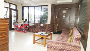 A reception area with a wooden reception desk, a waiting area with patterned sofas, and a large window providing a view of the outdoors. - Hotel Sachidanand (Rahata)