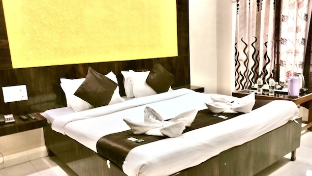 A neatly made bed with white linens and a black accent, a bedside table with a lamp, and a wall-mounted TV. - Hotel Sachidanand (Rahata)