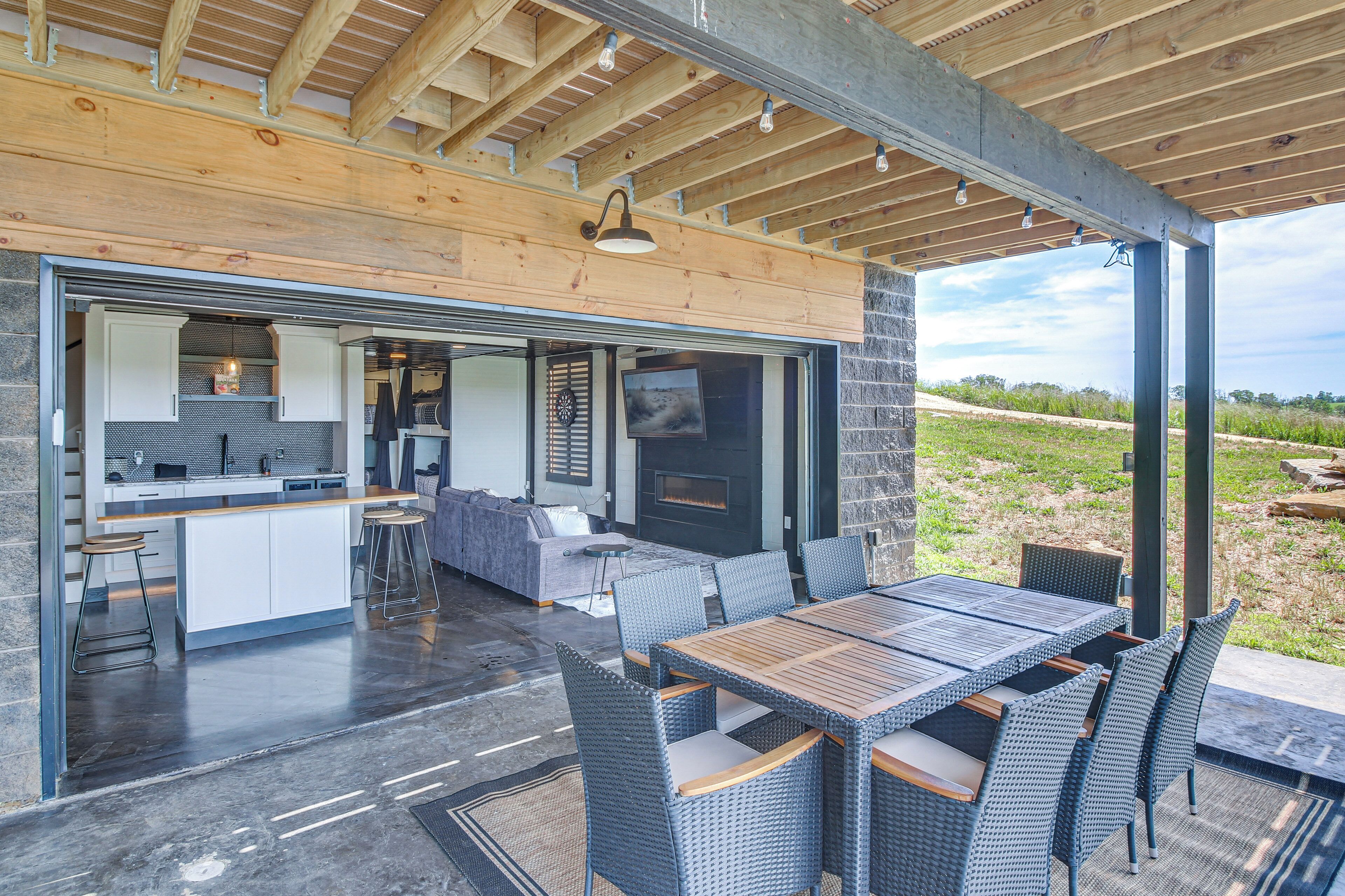 An open-plan kitchen and dining area with a stone wall, wooden ceiling beams, and a large window overlooking a grassy landscape.