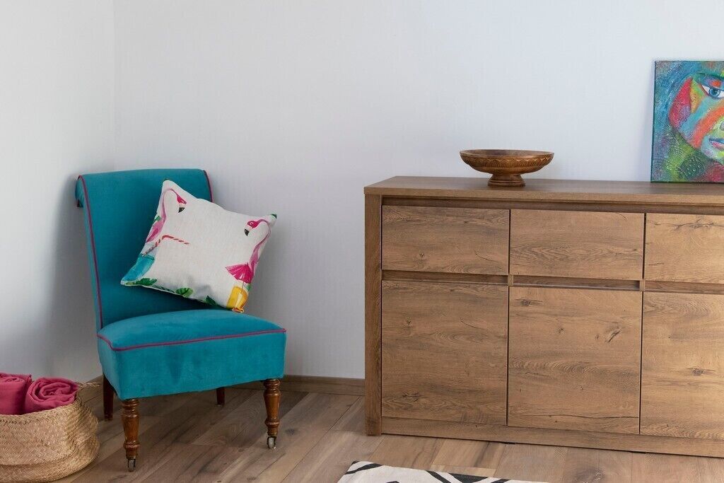 Un fauteuil turquoise agrémenté d’un coussin à motifs floraux, un meuble en bois sur lequel est posé un bol, et un tableau coloré accroché au mur.