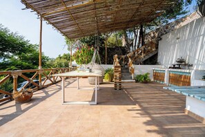 An outdoor terrace with a bamboo pergola, a table with chairs, a hanging plant, and a pool area.