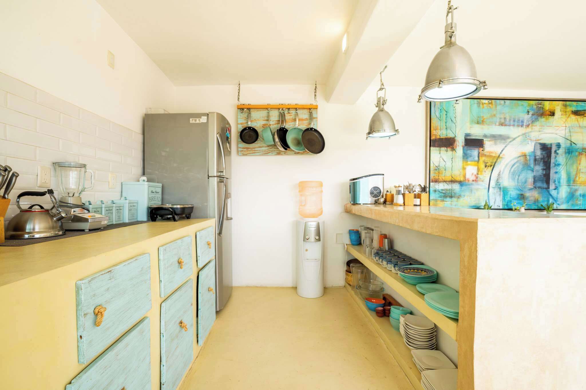 A compact kitchen with a refrigerator, stove, and various utensils. There is a water dispenser and a shelf with dishes.