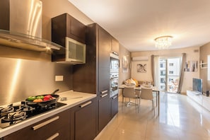 A modern kitchen with a stove, a pan with vegetables, and a dining area with a table and chairs. - Modern and Comfortable Apart Savoy Garden (Gzira)