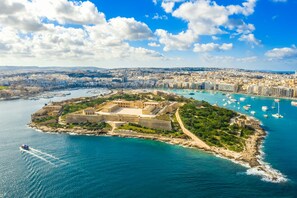 Vista aérea de una ciudad costera con una fortaleza destacada, un puerto deportivo con barcos y un cielo azul despejado.