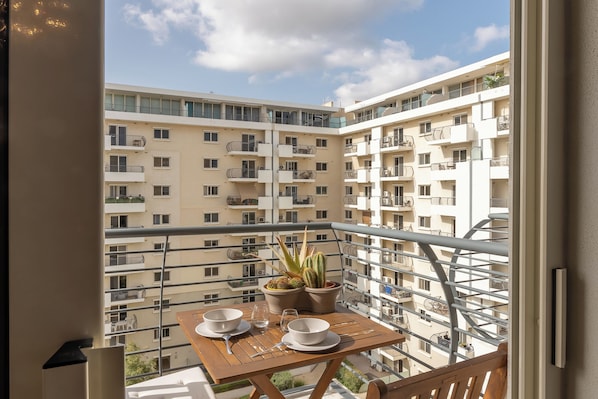 A balcony with a table set for two, potted plants, and a view of adjacent apartment buildings.
