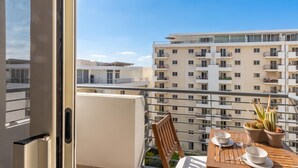 A balcony with a table set for two, a chair, a potted plant, and a view of a multi-story building. - Modern and Comfortable Apart Savoy Garden (Gzira)