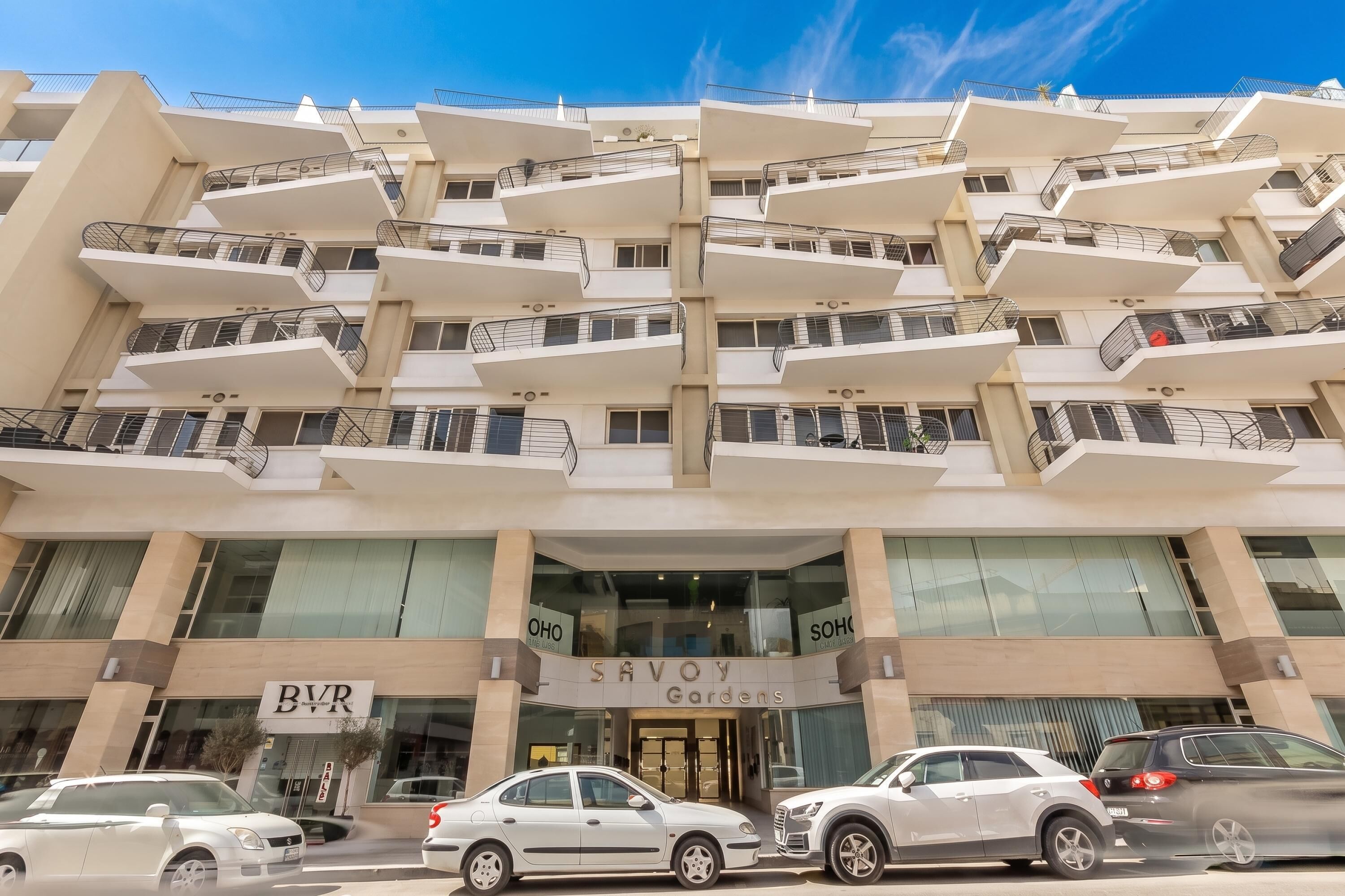 A multi-story building with a modern architectural design, featuring balconies on each floor, a prominent entrance with signage, and several parked cars in front.
