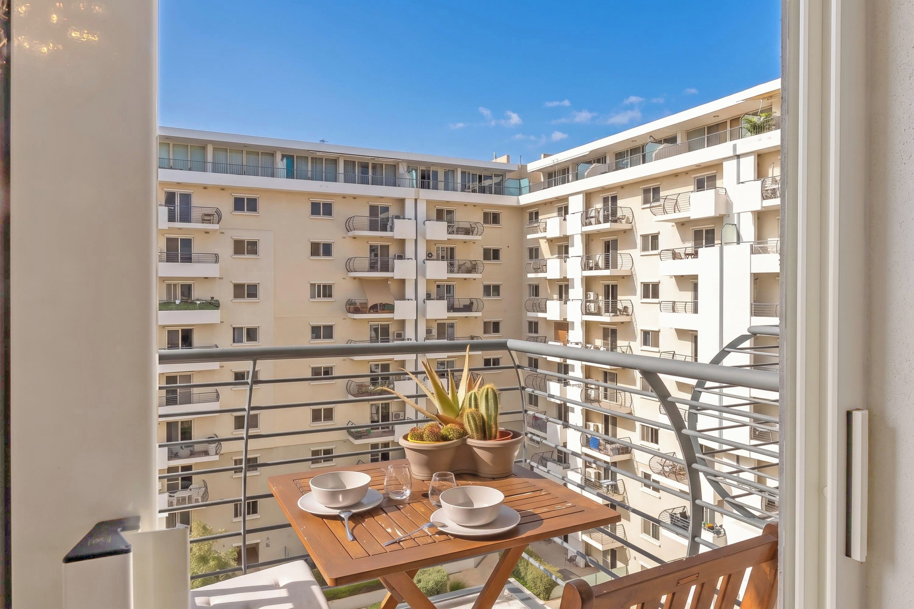 A balcony with a table set for two, potted plants, and a view of a multi-story residential building.