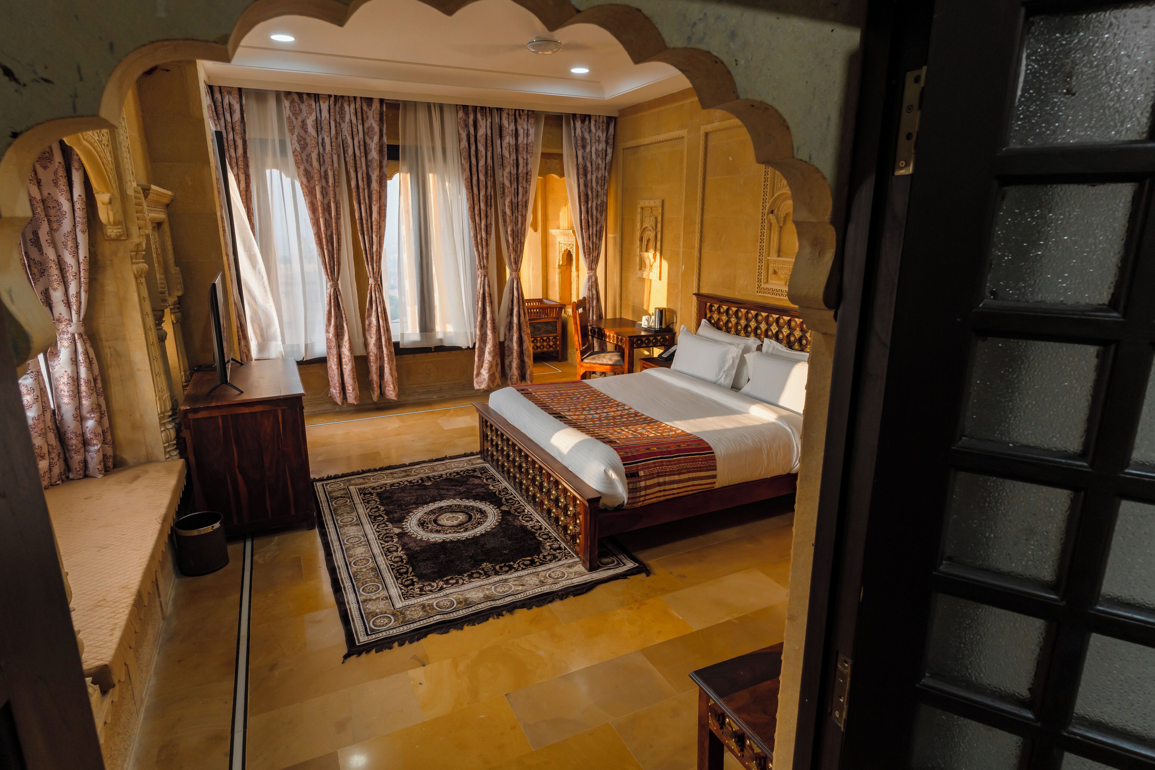 A hotel room with a large bed, a wooden nightstand, a desk with a chair, and a patterned rug.