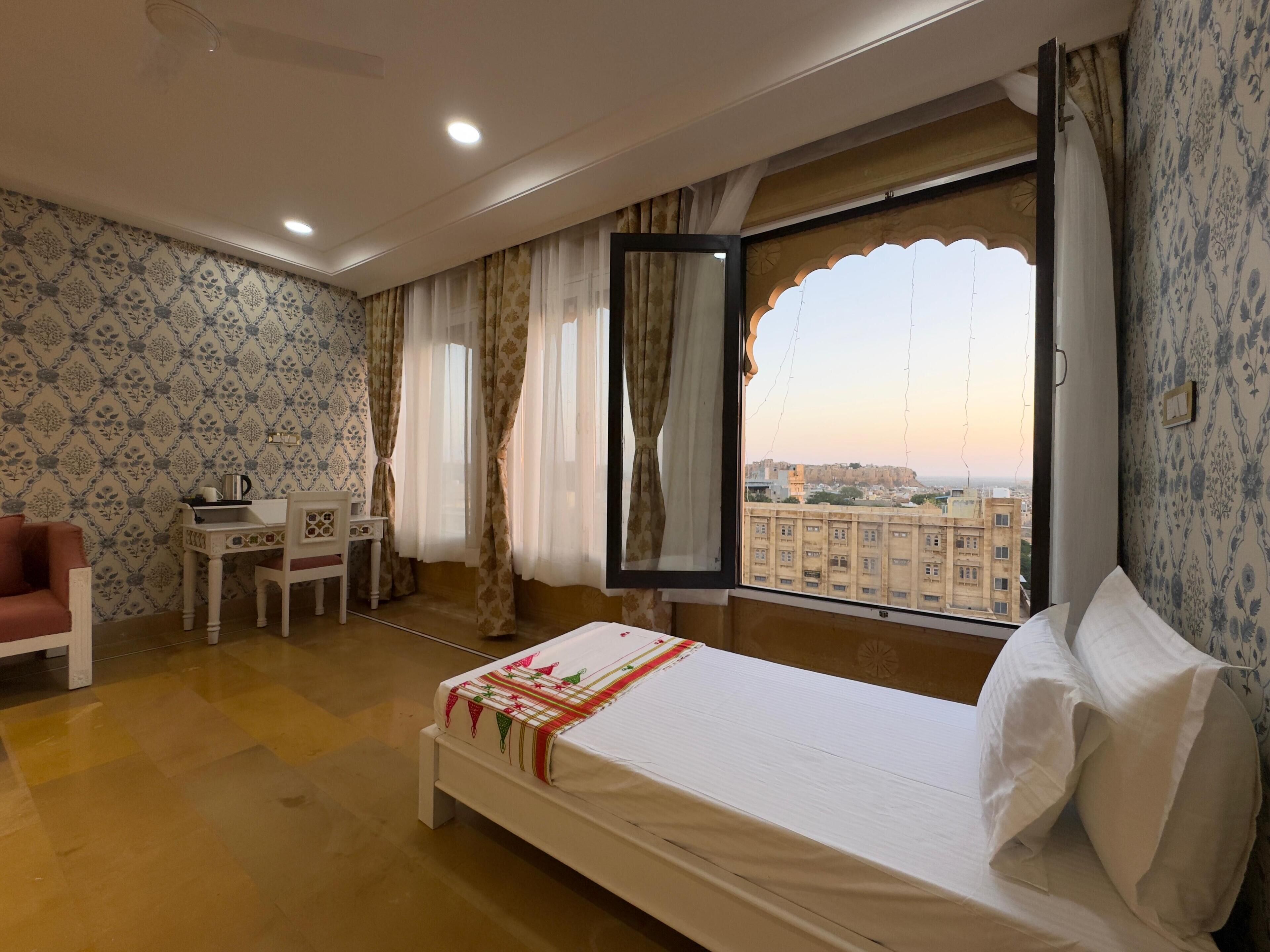 Habitación de hotel con cama, un pequeño escritorio con hervidor, una silla y vista de un paisaje urbano a través de una ventana grande con cortinas.