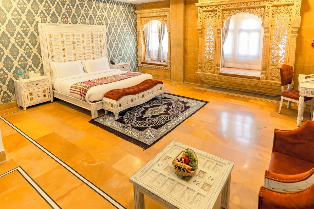 Une chambre d’hôtel avec un grand lit, une table de chevet, une petite table sur laquelle se trouve un panier de fruits, et une chaise.