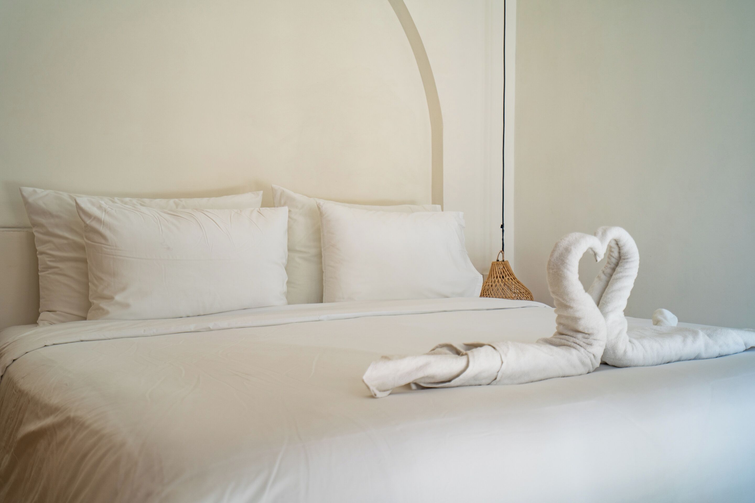 Una cama cuidadosamente arreglada con sábanas y almohadas blancas, una cesta tejida y una toalla doblada en forma de cisne.